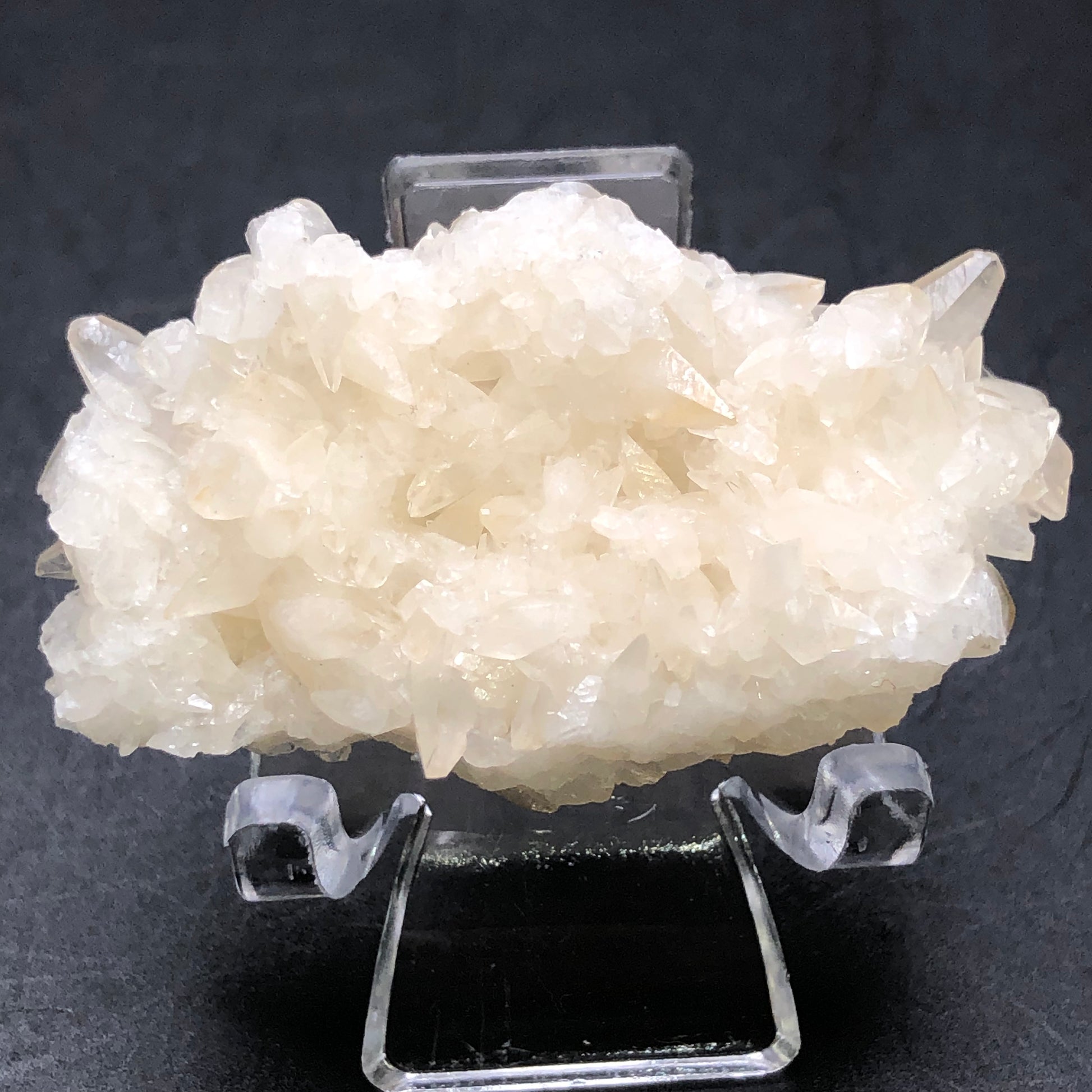 A stunning specimen of translucent, cream-colored crystals from The Crystalary rests on a clear stand against a dark background. Sharp and unevenly textured like peach calcite crystals from the Southwest Mine in Hendricks Gulch, Bisbee, Arizona, their natural elegance captivates.