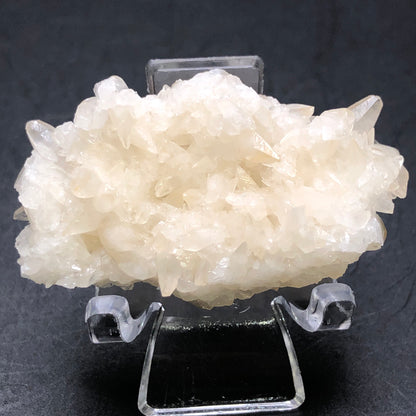 A stunning specimen of translucent, cream-colored crystals from The Crystalary rests on a clear stand against a dark background. Sharp and unevenly textured like peach calcite crystals from the Southwest Mine in Hendricks Gulch, Bisbee, Arizona, their natural elegance captivates.