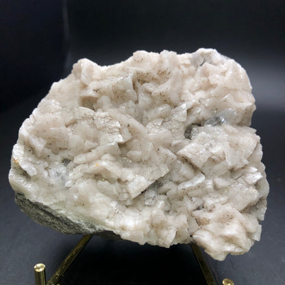 A close-up of the AUCTION - Dolomite & Calcite specimen from The Crystalary, showcasing dolomite crystals adorned with numerous white calcite formations against a dark background, elegantly displayed on a small stand. The surface has a rough and textured appearance.