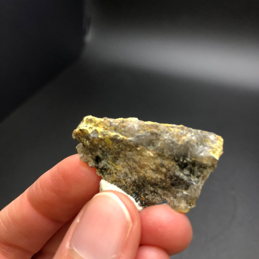 A hand holding a small, rough rock with a mix of gray and yellowish colors, reminiscent of the Pottsite from The Crystalary's auction at Linka Mine in the Spencer Hot Springs Mining District, Lander County, Nevada, USA, set against a dark background.