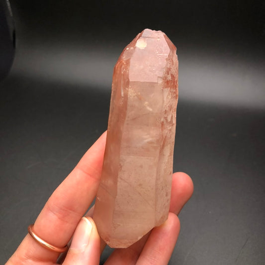 A hand holds a sizable, pointed piece of Pink Lemurian Quartz from The Crystalary's AUCTION collection against a dark backdrop. This semi-transparent crystal, originating from Minas Gerais, Brazil, features a subtle pink hue and rough edges that glisten gently. The person's fingers are decorated with a gold ring.