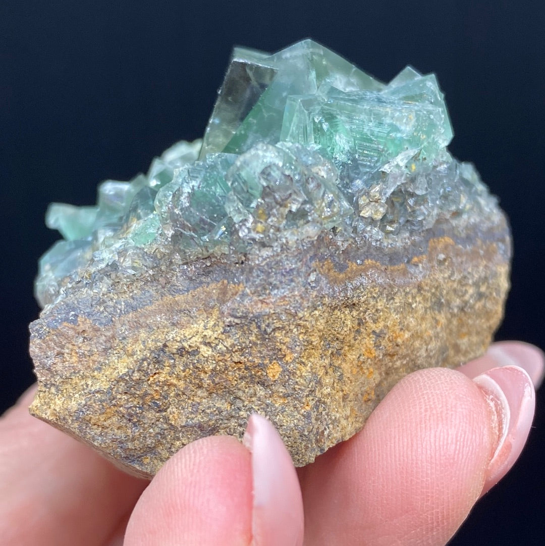 A hand holding a piece of Fluorite - Rainbow's End Pocket from The Crystalary, sourced from the Lady Annabella mine in Co Durham, UK. The rock has a rough, brownish base with green, translucent crystals protruding from its surface and displaying a geometric, angular structure against a solid dark background.