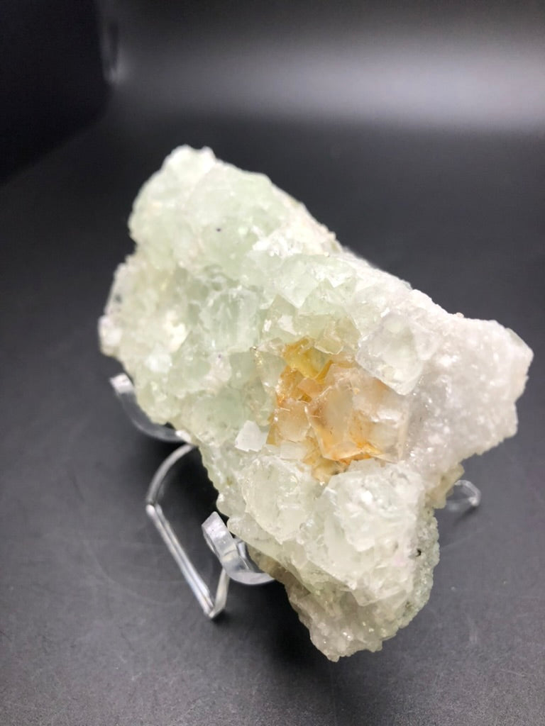 A significant specimen from The Crystalary features a large, pale green fluorite crystal with a rugged, textured surface placed on a small stand. At its core, vibrant yellowish-orange crystals sparkle like hidden gems. This unique piece originates from the renowned Xianghualing Mine in Linwu Co., Chenzhou, Hunan, China, and is beautifully accentuated by the rich background that highlights its exquisite details.