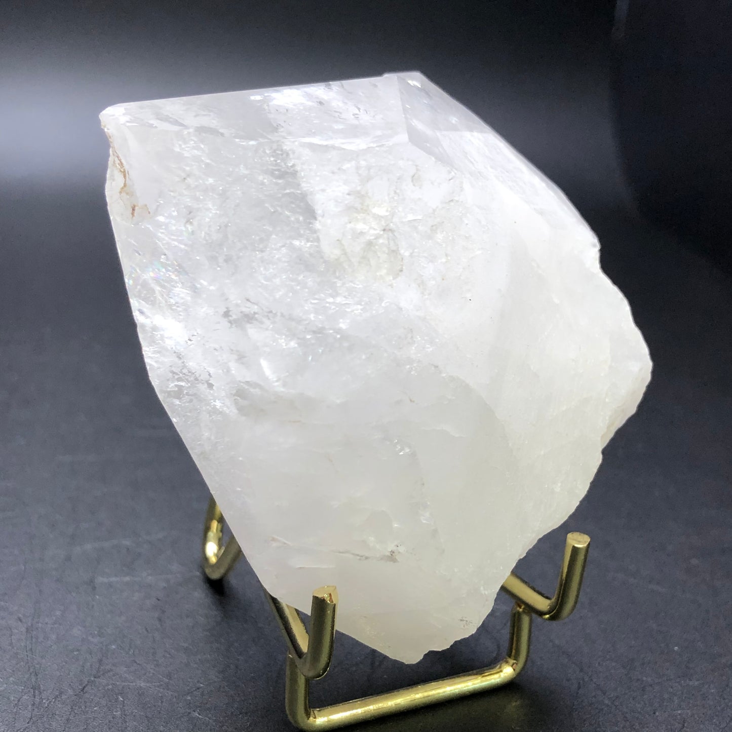 The Crystalary's AUCTION- Quartz- unk locality features a large, translucent quartz point with a rough surface that is elegantly displayed on a small metal stand against a dark background. Its irregular shape and milky white color highlight its natural transparency.