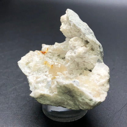 A textured piece from The Crystalary's AUCTION series, featuring a geode with calcite from Worthington, Ohio, USA, rests on a transparent stand against a dark background. The white and pale gray rock is reminiscent of a druzy geode and contains specks of orange hues within the crystal cluster, adding an unexpected warmth typical of honey calcite formations.