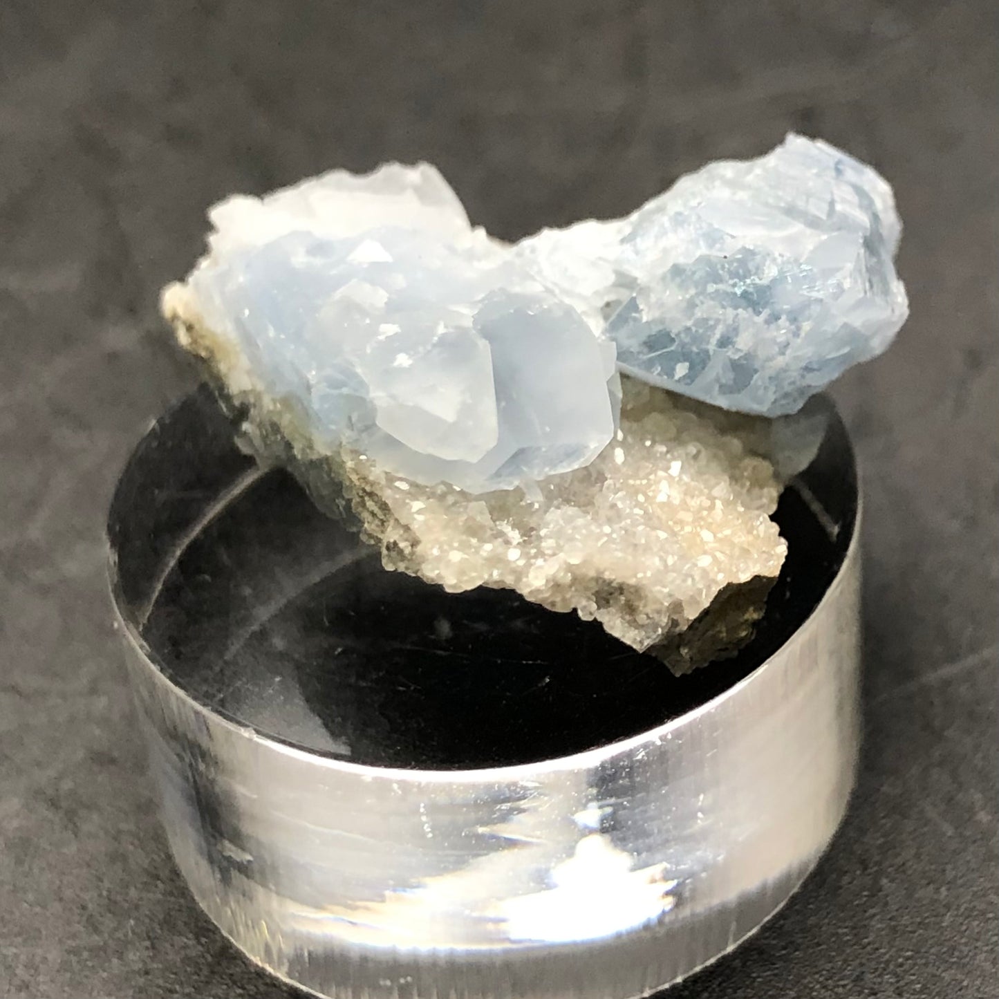 A collection of light blue Celestine crystals from Meckley's Quarry, crafted by The Crystalary, sits on a grayish mineral base and is showcased on a reflective cylindrical platform. These miniature gems feature a translucent texture that creates a striking contrast against the dark background.