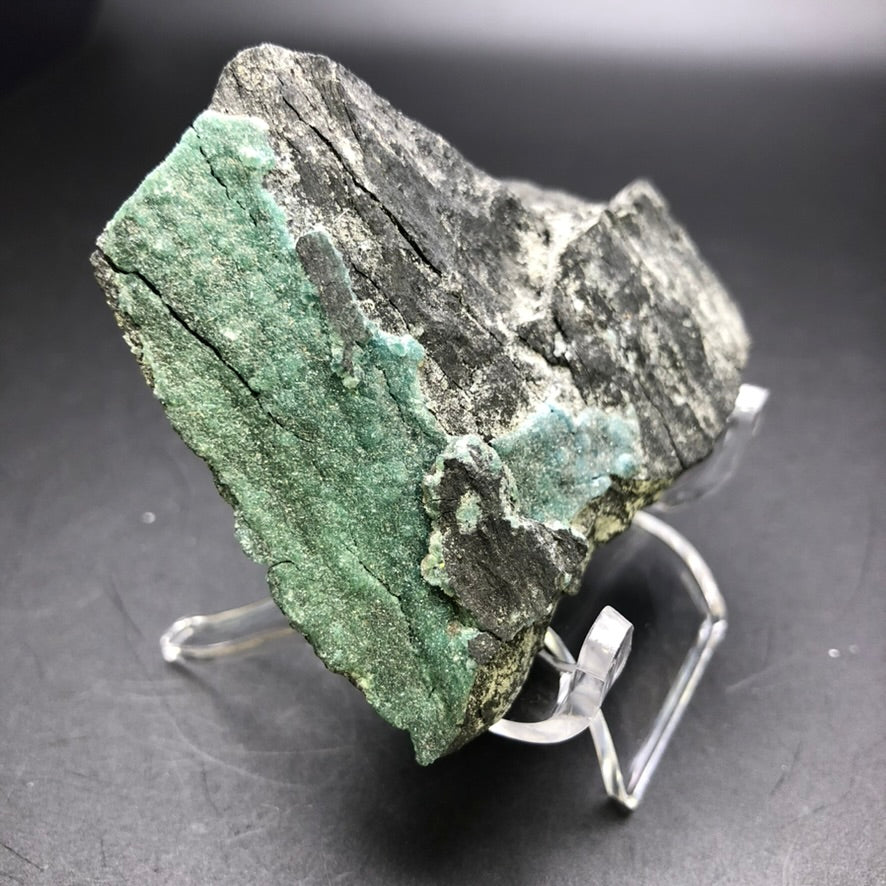 The mineral specimen, identified as "AUCTION- Wavellite" by The Crystalary from Ronneburg, Gera, Thuringia, Germany, showcases a rough and rocky texture with black and gray tones accented by a striking green crystalline section. This remarkable rock is elegantly presented on a clear plastic stand against a simple dark backdrop.