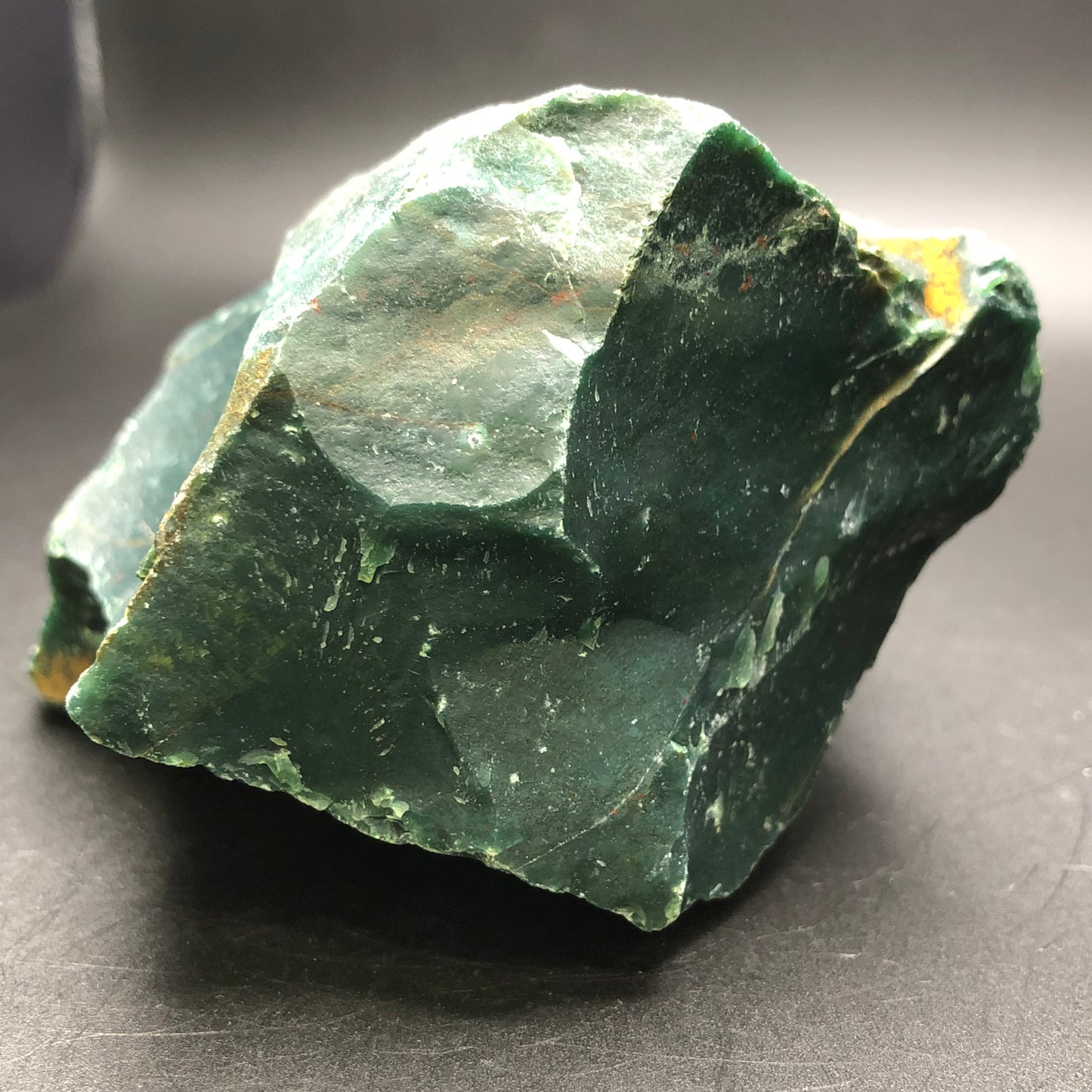 A rough, unpolished piece of AUCTION- Bloodstone- unk locality from The Crystalary displays smooth and jagged surfaces against a simple dark background.