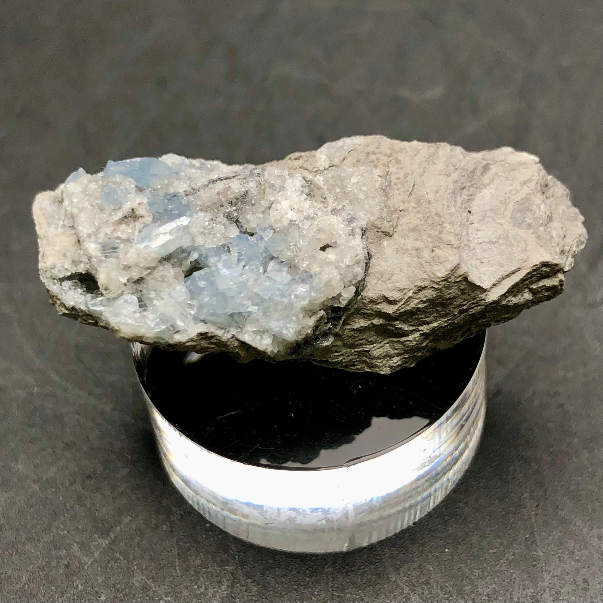 A close-up of a rough stone with bluish crystal formations is displayed on a round, clear stand against a dark background. Its appearance echoes that of the Celestine miniatures from Meckley's Quarry. The stone beautifully combines earthy and crystalline textures and is part of the "AUCTION- Celestine" collection by The Crystalary, originating from Meckley's Quarry, Mandata, Northumberland County, Pennsylvania, USA.