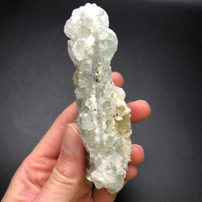 A hand displays the AUCTION- Fluorite from The Crystalary, showcasing an elongated, translucent mineral with cluster formations and fluorite cubes. This specimen, sourced from the Xianghualing Mine in Linwu Co., Chenzhou, Hunan, China, features a light green hue and a slightly rough texture against a dark background.