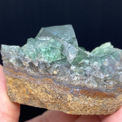 A hand holding a chunk of rock with green, translucent crystals from the Rainbow's End Pocket at the Lady Annabella mine in Co Durham, UK. The crystals sit atop a rough, brownish stone base and seem to form a jagged, uneven surface. The background is solid black, emphasizing the texture and color of this striking Fluorite crystalline formation from The Crystalary.