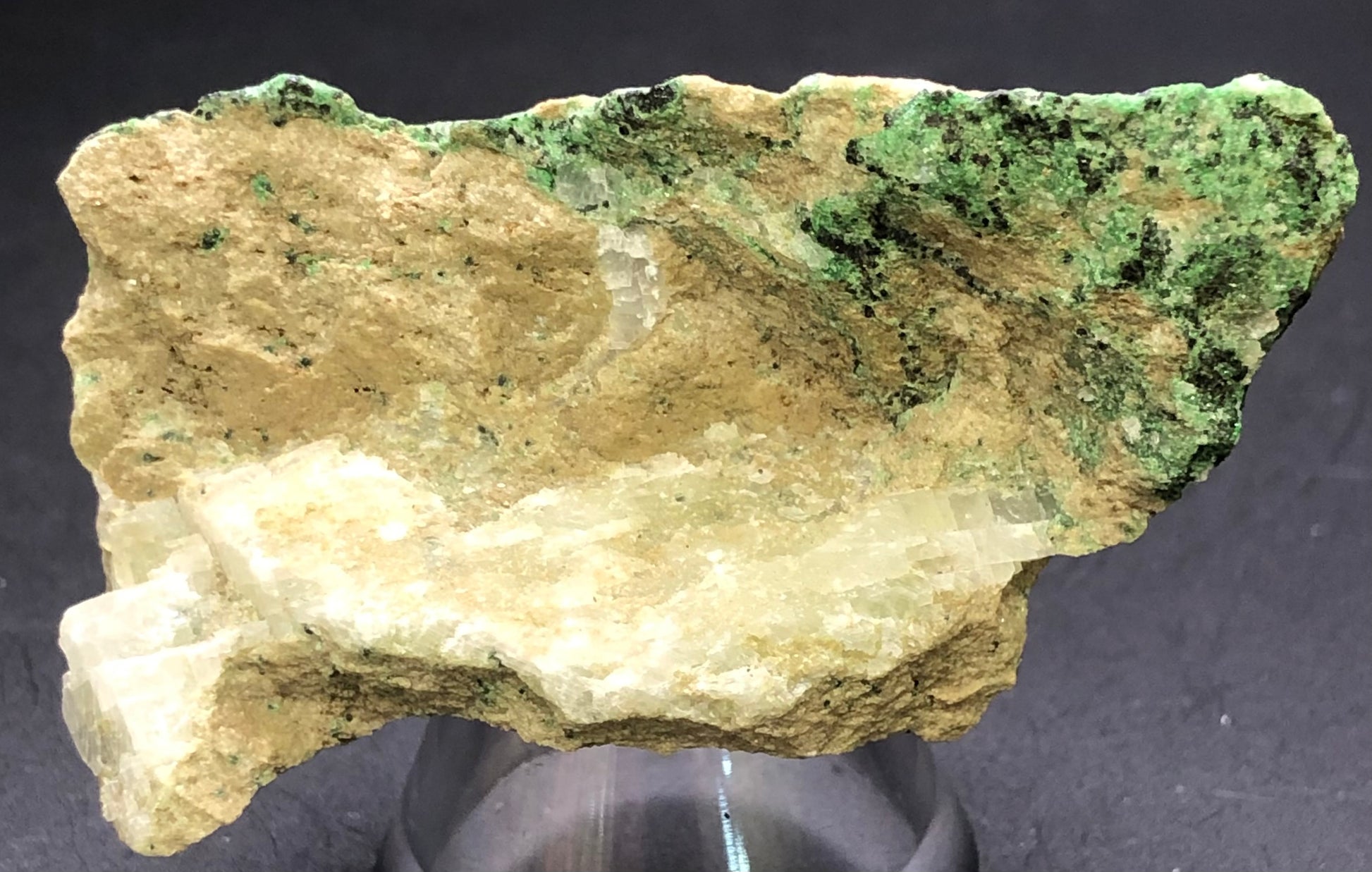 The Crystalary's auction item features an unpolished Uvarovite Garnet stone from Magog, Quebec, Canada. With its beige and green textured surface resembling garnet speckles, it rests on a small dark stand.