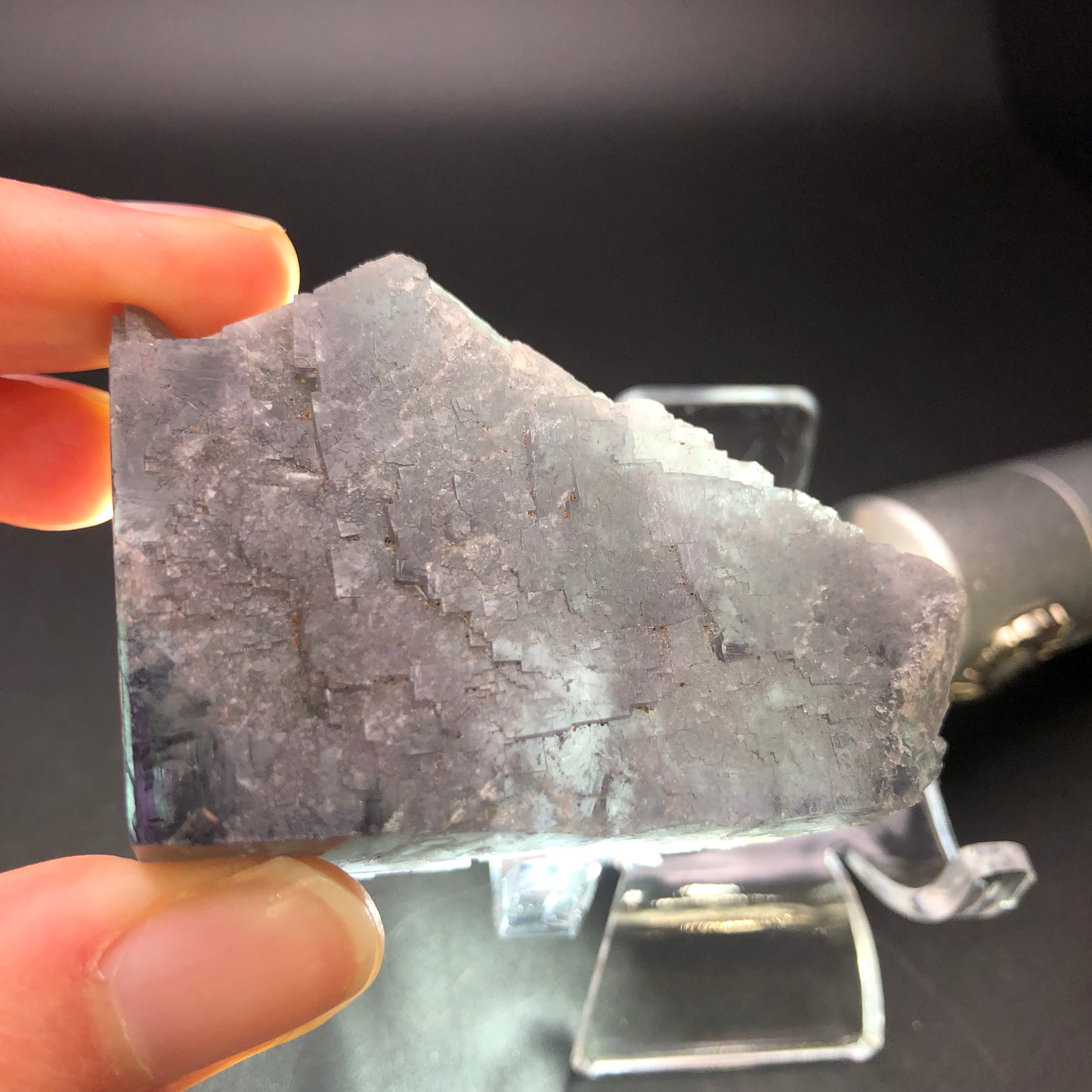 A close-up of a hand holding a triangular piece of translucent gray fluorite crystal, displayed on a stand, showcases its natural, rough texture that mirrors discoveries from the Lady Annabella Mine. Its internal structures and hues capture the enigmatic charm of purple-black cubes. The dark background provides a striking contrast. This piece is part of The Crystalary's AUCTION collection: Fluorite- Purple Rain Pocket, sourced from Lady Annabella Mine in Eastgate, Weardale, Co. Durham, England.