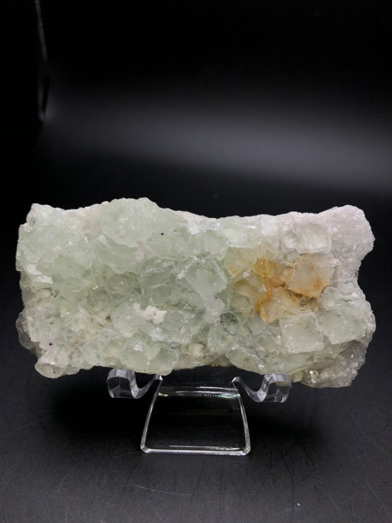 Displayed on a clear stand, a light green piece from The Crystalary, showcasing raw calcite with orange-brown inclusions against a black background. This specimen originates from the Xianghualing Mine in Linwu County, Chenzhou, Hunan, China. It features rough crystalline structures and a translucent appearance that evokes the presence of embedded fluorite cubes.