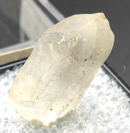 A close-up of the AUCTION- Selenite from Willow Creek, Nanton, Alberta, Canada by The Crystalary showcases its translucent nature and slightly yellowish hue. Positioned on a textured white surface, this angular-shaped crystal features visible imperfections and a glassy transparency with small brown inclusions.