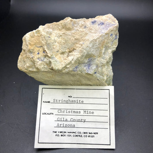 The Stringhamite mineral sample from The Crystalary is showcased with a label emphasizing its origin from the famous Christmas Mine in Gila County, Arizona.