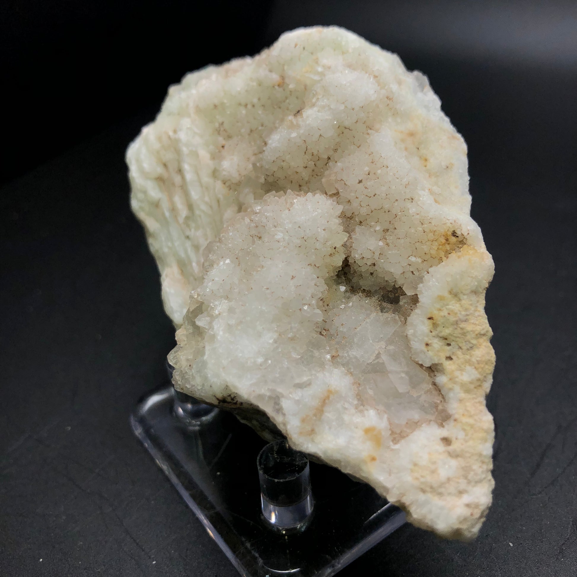 A close-up of the AUCTION - Calcite, Quartz piece by The Crystalary, showcasing a rough druzy white quartz crystal cluster with numerous small, shimmering facets. The irregularly shaped mineral is presented on a clear plastic stand against a dark backdrop.