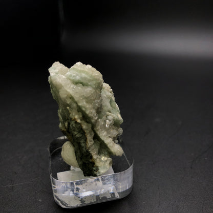 A stunning mineral specimen, featuring rough and jagged green and white crystals from D&G Quarry, is elegantly displayed on a clear acrylic stand against a dark backdrop. The Apophyllite on Prehnite from Woodbury, Litchfield County, Connecticut, USA shimmers with its raw textures, showcasing the natural beauty of its formation. This exquisite piece is part of The Crystalary's auction collection.