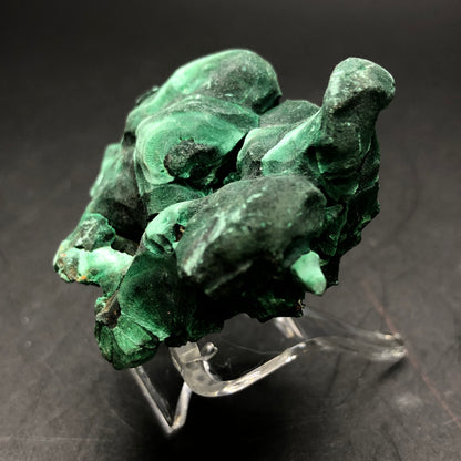 A raw malachite specimen from the Liufengshan Mine in Anhui, China, presented by The Crystalary, showcases swirling patterns and is elegantly displayed on a transparent acrylic stand set against a dark background.