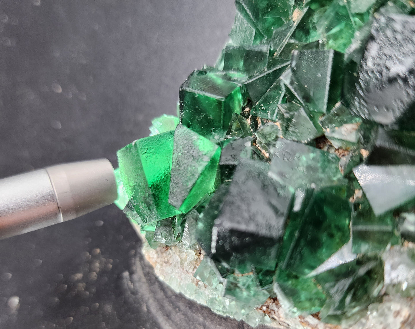 Close-up of a cluster of green cubic fluorite crystals from The Crystalary's Fluorite- Greedy Hog Pocket, Diana Maria, Co. Durham, England on a grey background. A silver pointer touches one of the transparent green crystals, highlighting its clarity and geometric shape. The crystals exhibit a vibrant and deep green hue.