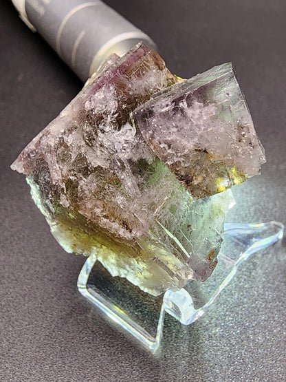 A close-up photograph of a specimen from The Crystalary's Fluorite - Purple Haze Pocket, Diana Maria Mine, Rogerley, Weardale, England showcases its multicolored hues of purple, green, and brown. The crystalline mineral is displayed on a translucent stand against a black background, with part of a cylindrical object visible in the background.