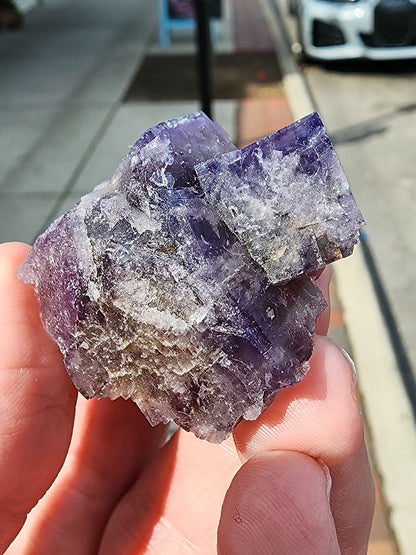 A hand holds a rough, purple and white mineral rock against a blurred street background with sidewalks and parked cars. The Fluorite- Purple Haze Pocket from the Diana Maria Mine, Rogerley, Weardale, England offered by The Crystalary showcases a crystalline structure with distinct, angular facets reminiscent of renowned finds from this region.