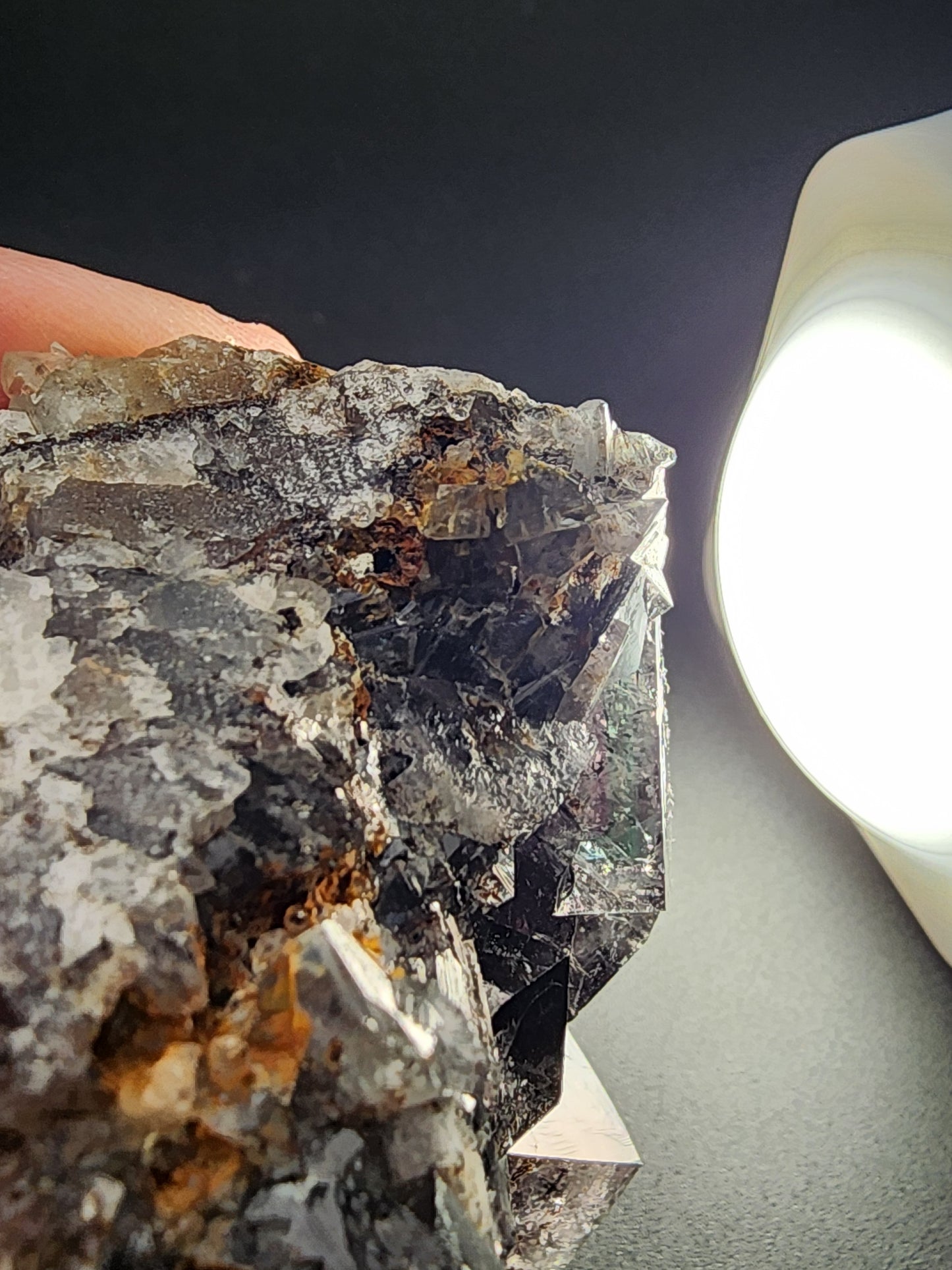 Close-up of a dark, textured Fluorite cluster from the Yum Yum Pocket, Diana Maria Mine, Frosterley, Weardale, England by The Crystalary. Held against a black background, it showcases clear and dark crystalline formations with vibrant daylight fluorescence. A bright, circular light source on the right illuminates parts of the minerals, creating reflective highlights.