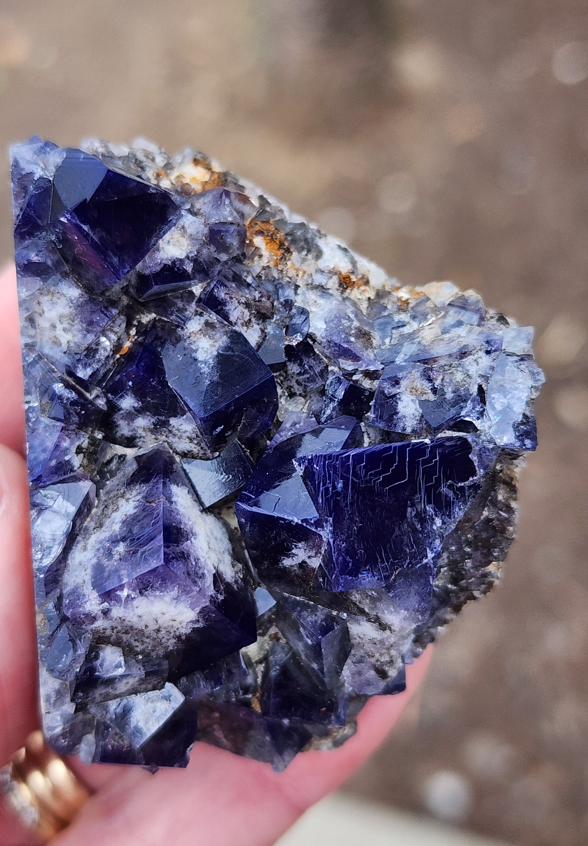 A close-up shot showcases a person's hand holding a piece of the Fluorite - Yum Yum Pocket, Diana Maria Mine, Frosterley, Weardale, England by The Crystalary. This deep purple and dark blue crystal mineral features a rough surface with sharp, geometric facets that reflect light beautifully, along with small areas of earthy matrix visible on its edges. The background is softly blurred.