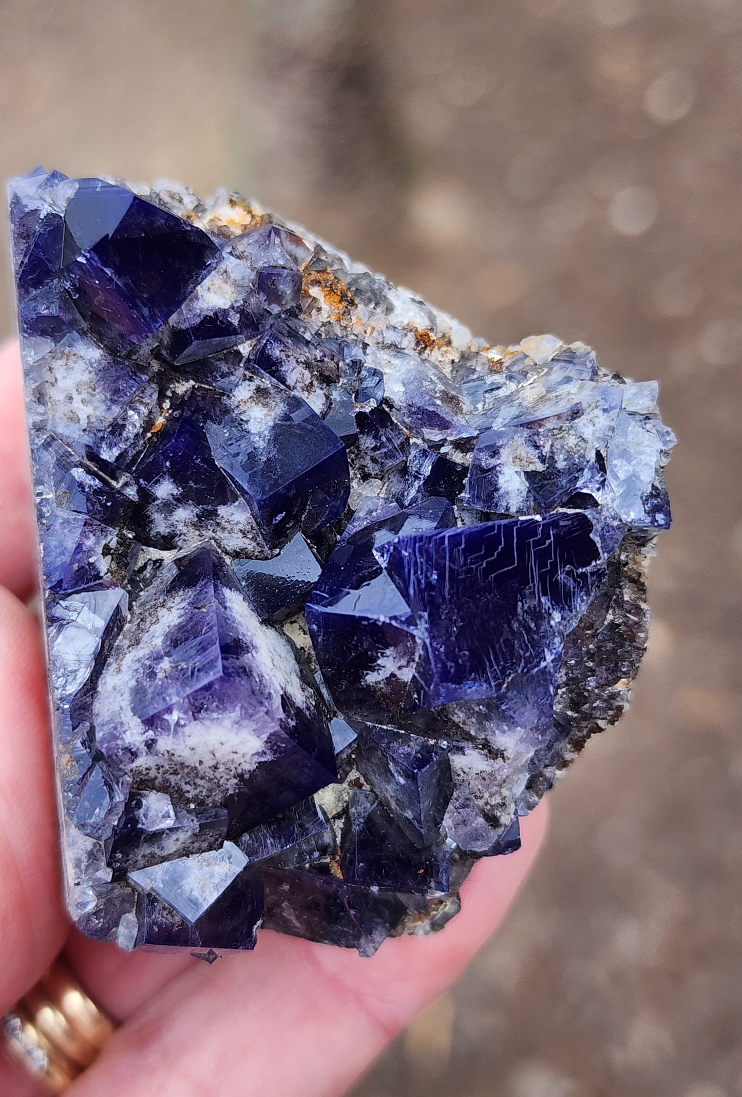 Close-up of a hand holding a piece of raw, dark purple gemstone with a crystalline structure. The Fluorite - Yum Yum Pocket from the Diana Maria Mine in Frosterley, Weardale, England features multiple faceted points and a rough, unpolished surface, contrasting against the blurred earthy background. This stunning cluster from The Crystalary exhibits daylight fluorescence.