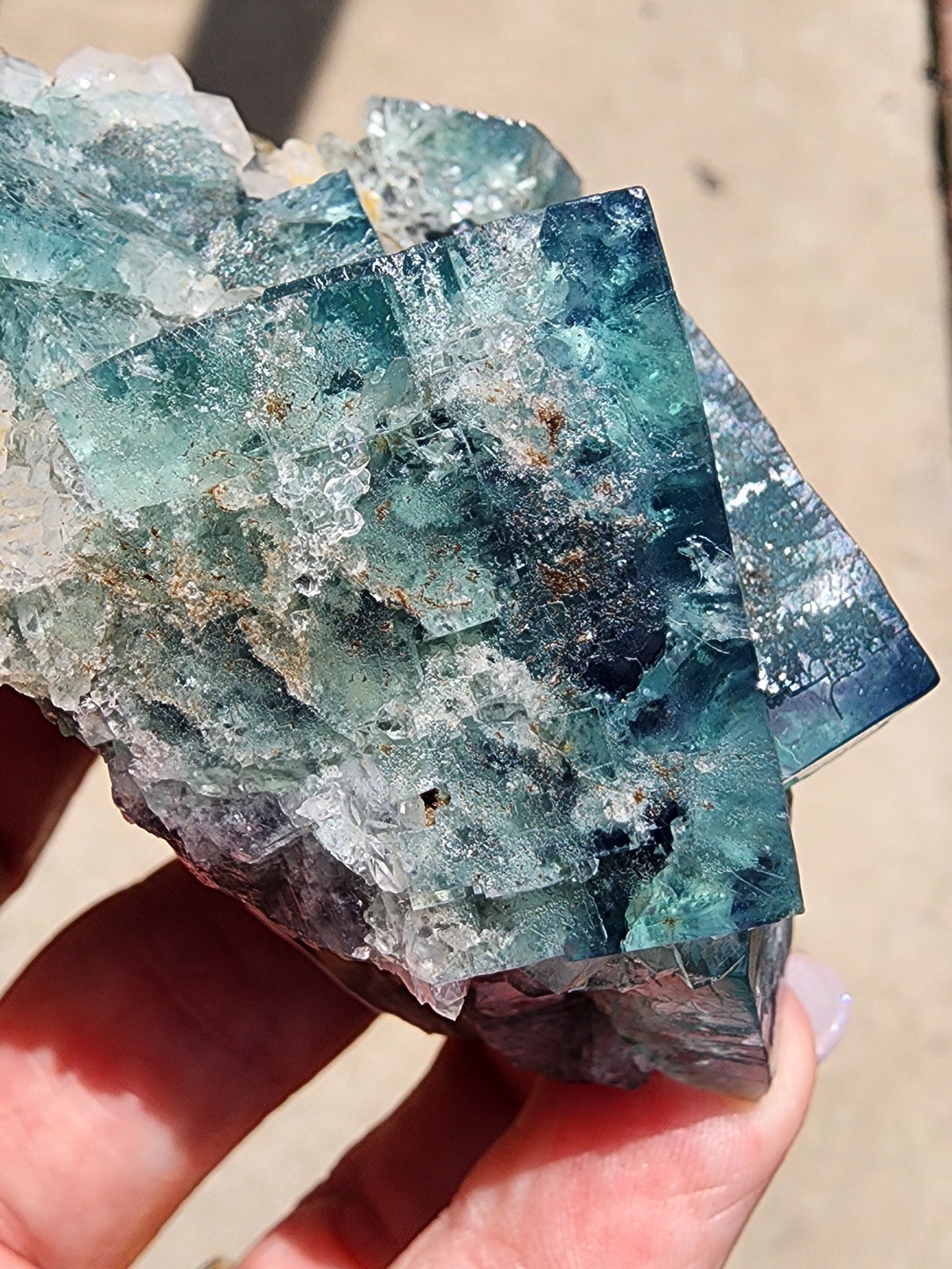 Close-up image of a hand holding a piece of raw Fluorite- Supernova Pocket, Diana Marie Mine, Weardale, Co. Durham, England from The Crystalary. The fluorite exhibits varying shades of blue, teal, and green with intricate translucent facets and natural textures. It almost looks like it could have come from a supernova pocket. The background is blurred, emphasizing the detail of the crystal.