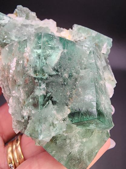 A close-up image shows a cluster of green Fluorite - Supernova Pocket from the Diana Marie Mine in Weardale, Co. Durham, England, held in a person's hand. The translucent crystals, with visible inclusions and varying shades of green, look like treasures from another world. The hand adorned with three gold rings contrasts beautifully against the dark background. This stunning specimen is brought to you by The Crystalary.