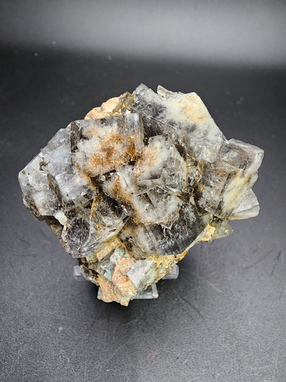 The Fluorite-Milky Way Pocket from The Crystalary, sourced from the Diana Maria Mine in Weardale, Co Durham, UK, displays an intricate cluster of raw mineral formations with cubic shapes and varying shades of gray, white, and brown. It sits against a plain black background, with some translucent sections subtly reflecting light in a manner reminiscent of earthy hues capturing the essence of a Milky Way Pocket.