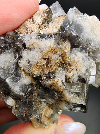 A close-up view of a hand holding a piece of mineral rock. The rock, likely the Fluorite-Milky Way Pocket from Diana Maria Mine, Weardale, Co Durham, UK by The Crystalary, features a combination of transparent and opaque crystal formations with white, brown, and grey colors. It reveals intricate, layered textures with a slightly cloudy appearance.