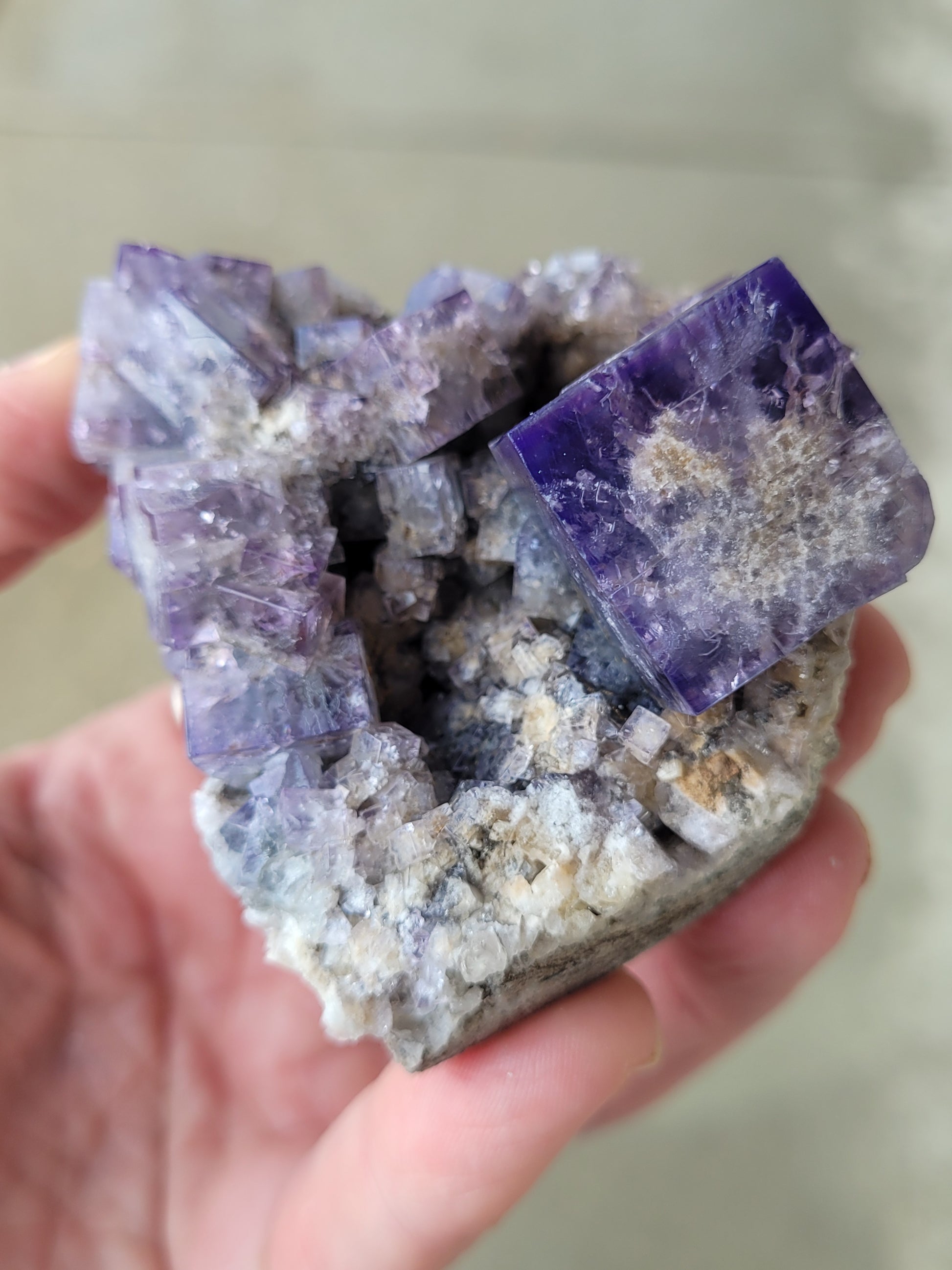 A hand holds a cluster of Fluorite- Purple Rain Pocket crystals from The Crystalary, sourced from the Fairy Hole Vein at Lady Annabella Mine in Eastgate, Weardale, Co. Durham, England. The crystals display sharp edges with deep inky purple and translucent colors. These intergrown, interpenetrant twins sit atop a matrix of contrasting lighter, earthy tones.