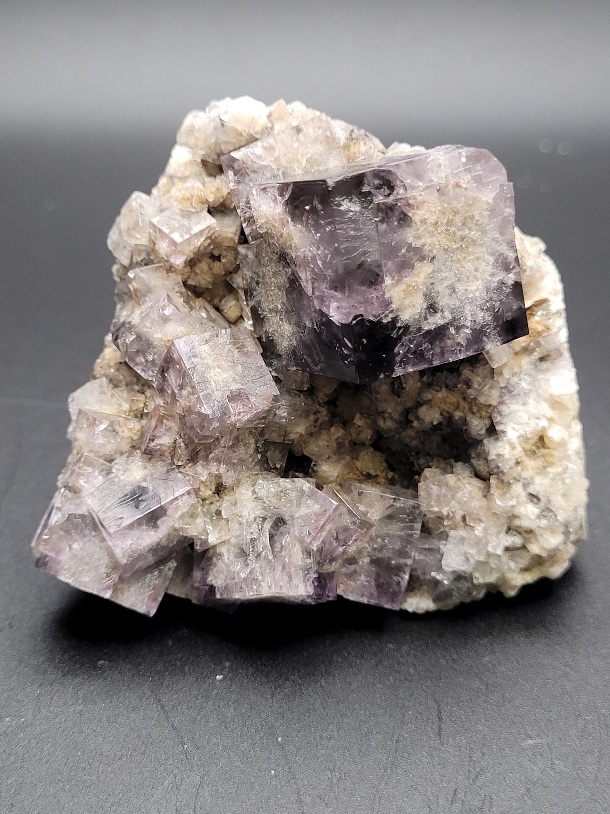 A close-up image of The Crystalary's Fluorite from the Purple Rain Pocket, Fairy Hole Vein, Lady Annabella Mine in Eastgate, Weardale, Co. Durham, England features cubic purple and translucent crystals. Some of these crystals include interpenetrant twins and are embedded in a rough white and tan matrix. The fluorite crystals exhibit a geometric, well-defined shape with deep inky purple hues and rest on a black surface.
