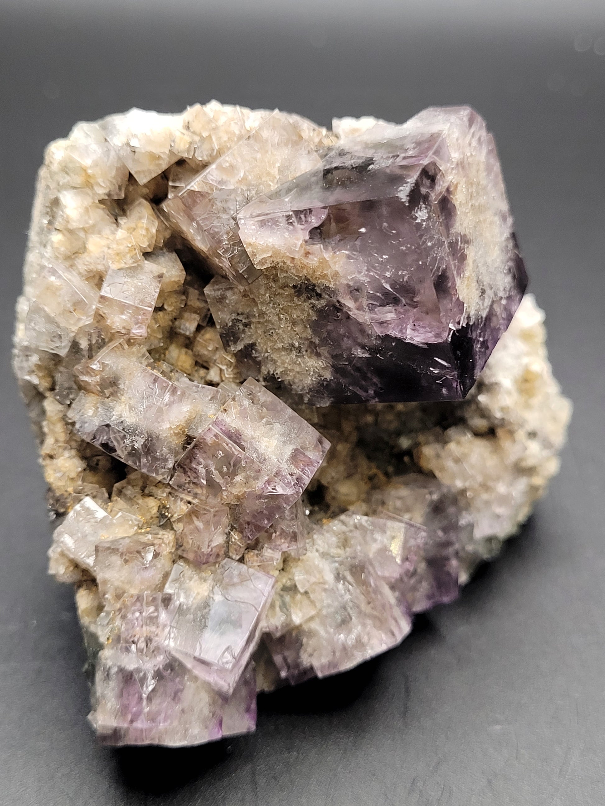 A cluster of cubic, translucent crystals with light purple and gray hues sits against a solid black background. The formations show distinct edges and varying sizes, with a rough, mineral base supporting the crystals. The reflective surfaces glisten like Fluorite- Purple Rain Pocket from the Fairy Hole Vein at Lady Annabella Mine in Eastgate, Weardale, Co. Durham, England by The Crystalary.