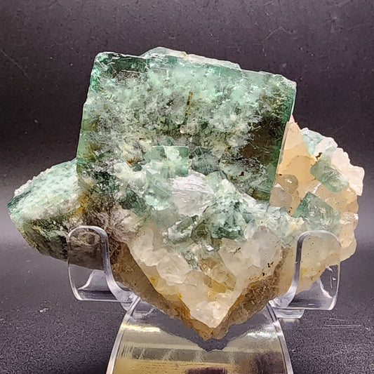 The Fluorite- Supernova Pocket from The Crystalary, sourced from the Diana Marie Mine in Weardale, Co. Durham, England, features a cluster of green fluorite crystals with slightly translucent edges and white crystalline formations interspersed throughout. This jagged structure sits on a stand against a dark background, its vibrant green hues and intricate details enhanced by the subtle daylight fluorescence.