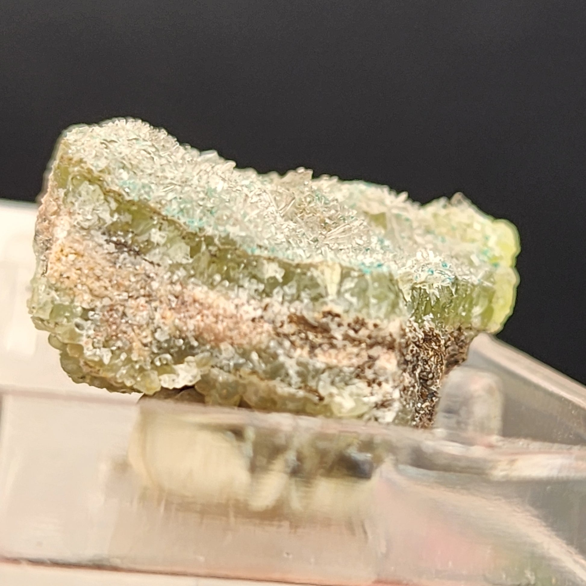 A close-up photograph of The Crystalary's AUCTION Hemimorphite, sourced from the 79 Mine in Dripping Spring Mountains, Gila County, Arizona, USA. The green and white hemimorphite mineral specimen exhibits a jagged texture with small reflective surfaces and is elegantly displayed on a clear stand against a blurred dark background.