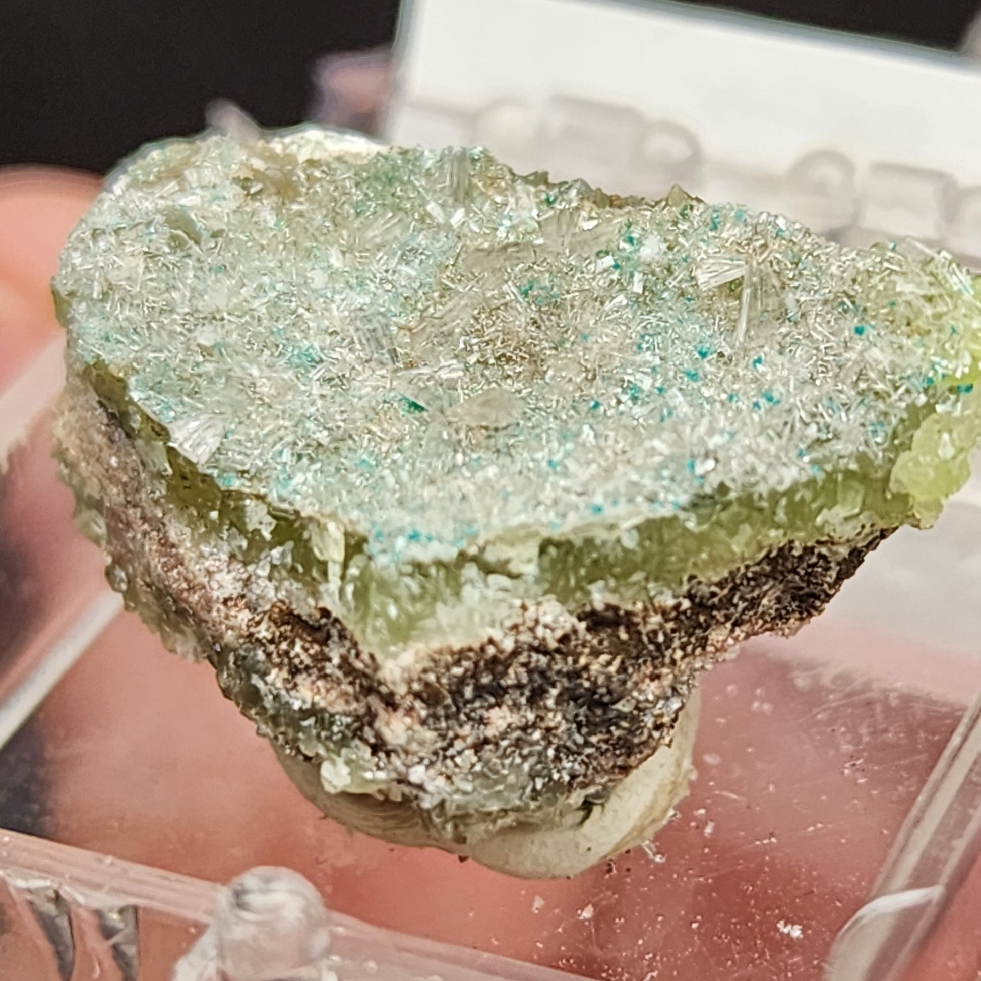 A close-up of the Hemimorphite specimen from the 79 Mine, available at The Crystalary, showcases a rough chunk of rock with a sparkling green surface. The rock structure reveals multiple layers, including a green crystalline top, a darker middle layer, and a lighter, almost yellowish base.