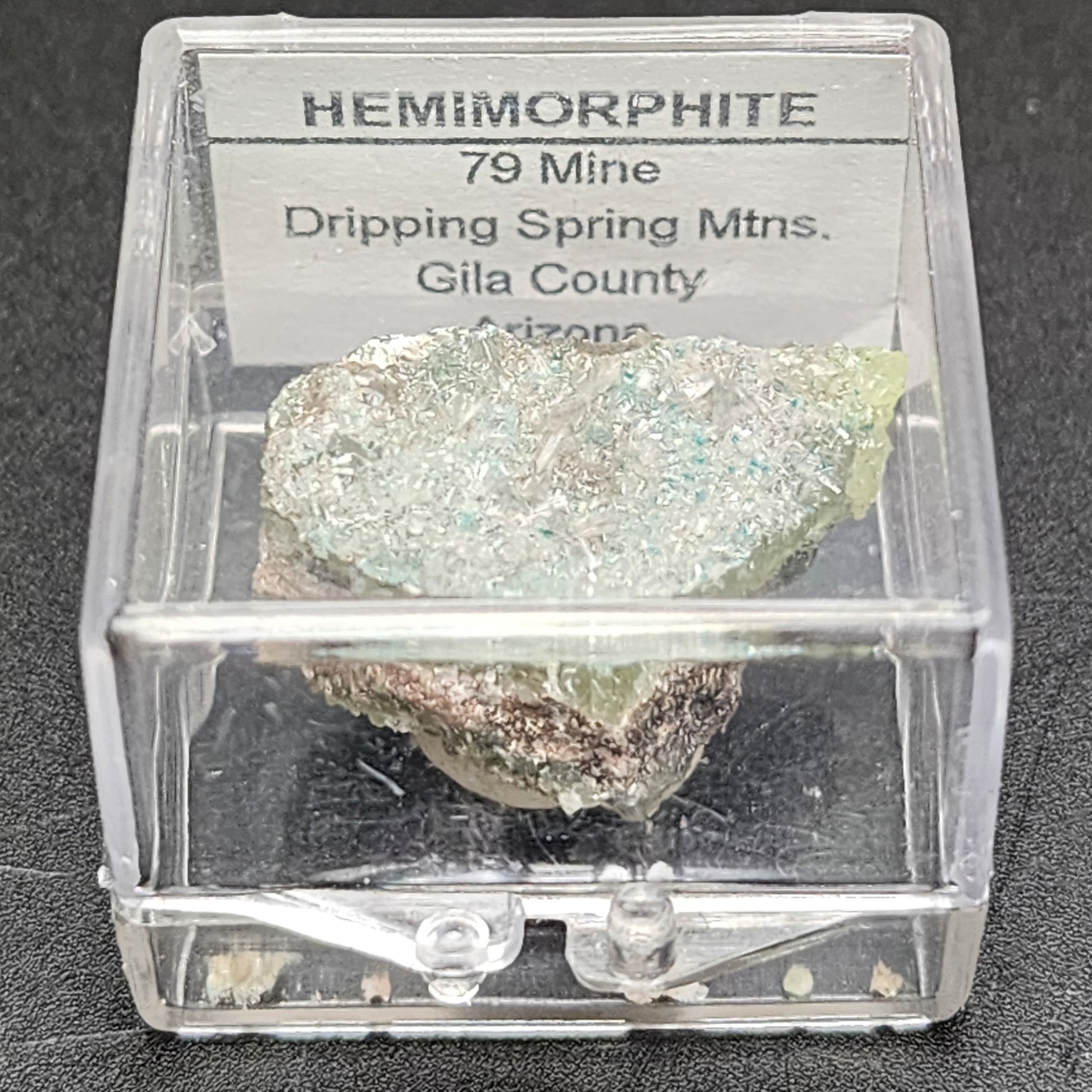 Housed in a clear plastic box, The Crystalary's stunning mineral specimen of hemimorphite is labeled "AUCTION Hemimorphite- 79 Mine, Dripping Spring Mtns., Gila County, Arizona, USA." This hemimorphite exhibits pale green and blue hues with a captivating crystalline texture.