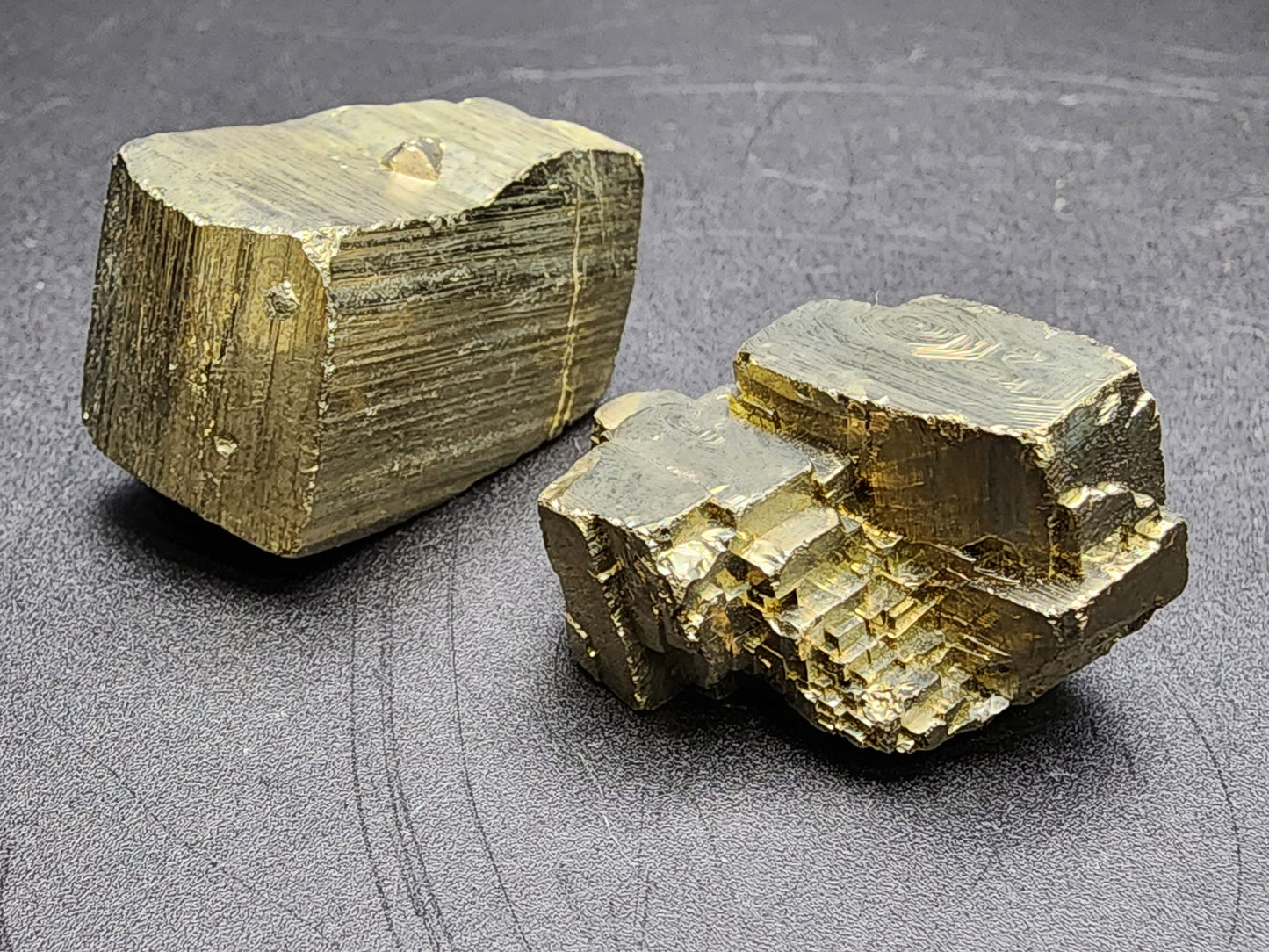 Two rough pyrite specimens from The Crystalary's AUCTION Pyrite collection, also known as fool's gold, are set against a black background. Sourced from La Rioja's renowned Ambas Aguas region, specifically Muro de Aguas in Spain, the crystal on the left displays a smooth, rectangular shape with distinct striations, while the one on the right has more irregular and blocky cubic formations.