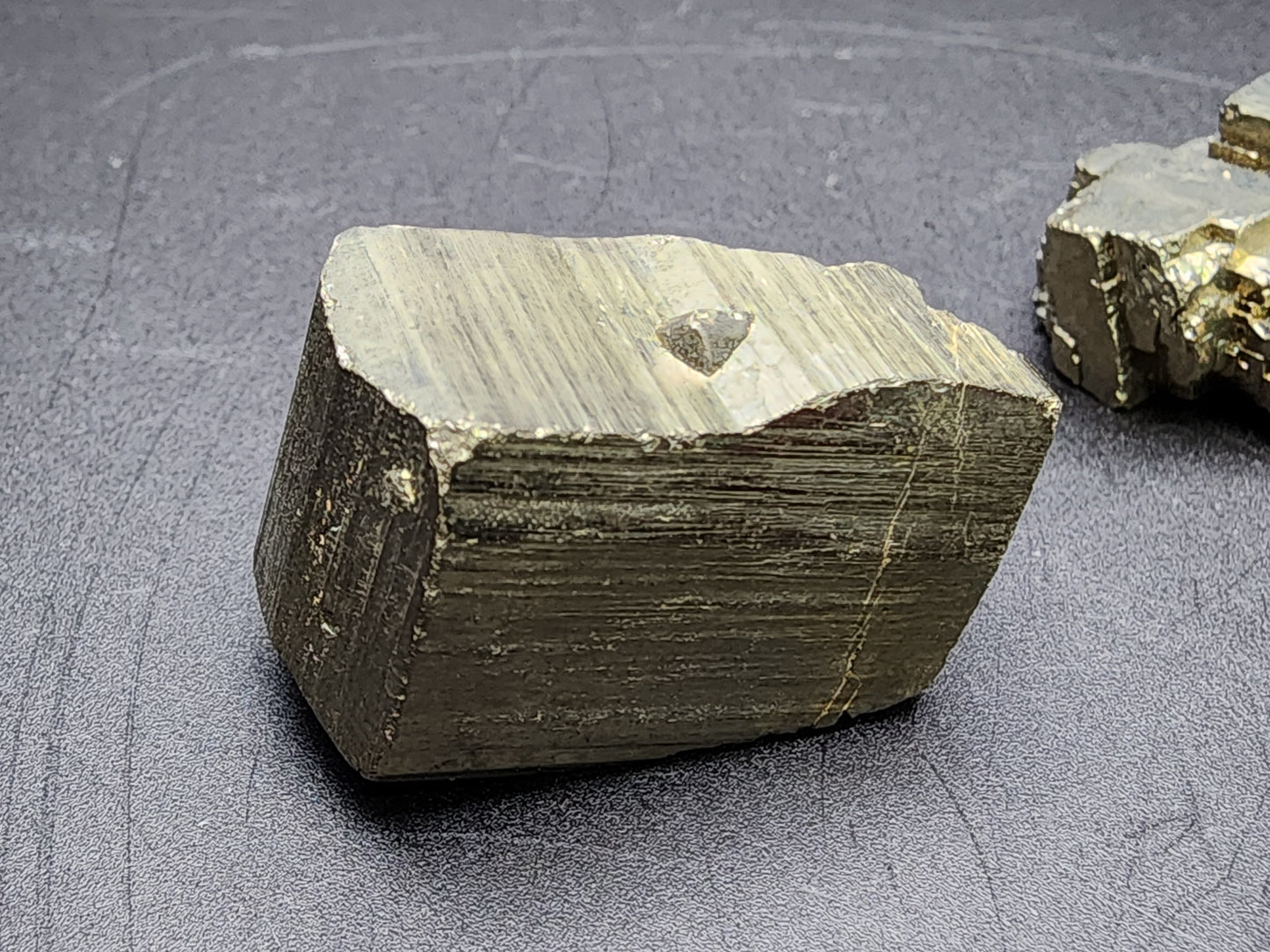 A small, rough piece of AUCTION Pyrite from Ambas Aguas, Muro de Aguas in La Rioja, Spain, also known as fool's gold, rests on a dark, textured surface. The mineral from The Crystalary exhibits a metallic luster and striated cubic structure with its characteristic grayish-gold color. Another specimen is partially visible in the background.