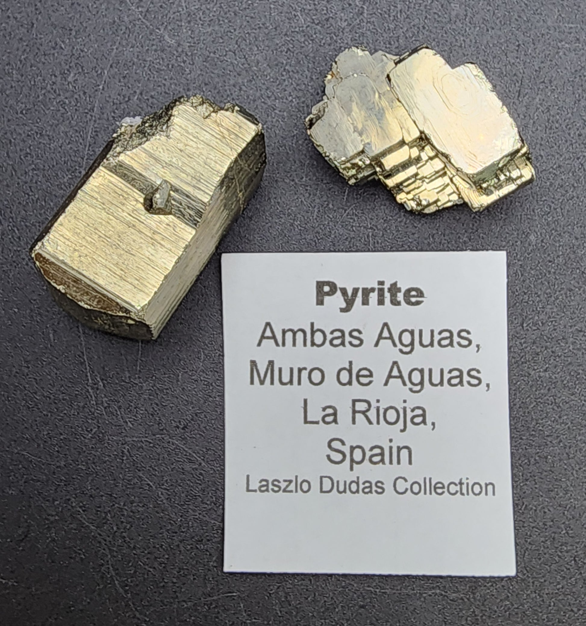 Two pyrite specimens from La Rioja, Spain, showcased on a dark surface beside a label that reads, "Pyrite - Ambas Aguas, Muro de Aguas, La Rioja, Spain," from the Laszlo Dudas Collection. Featuring metallic luster and distinct angular faces, they are part of The Crystalary's AUCTION series.