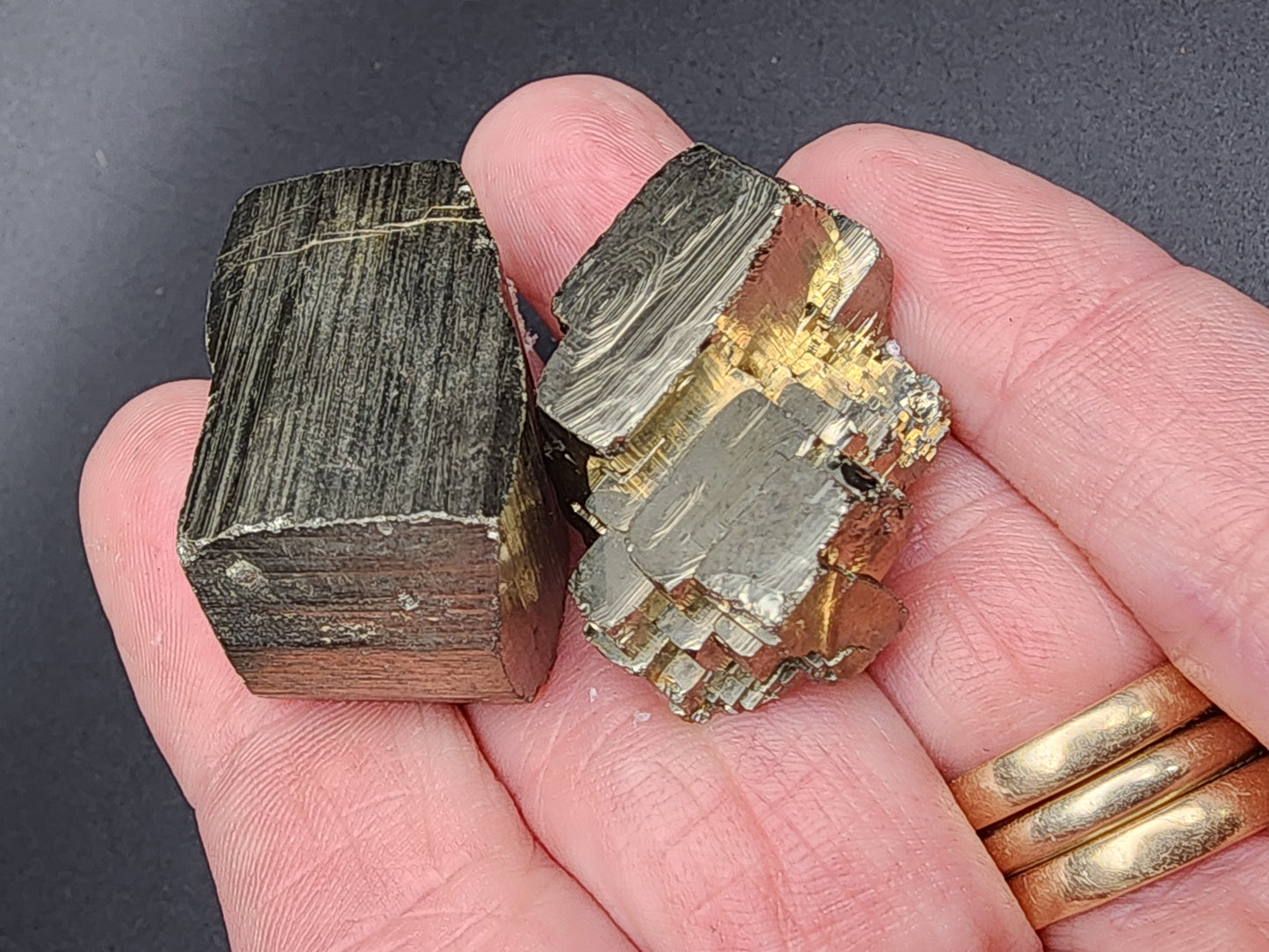 In Ambas Aguas, Muro de Aguas, La Rioja, Spain, a hand is holding two specimens of AUCTION Pyrite from The Crystalary. The specimen on the left is cubic and metallic with distinct striations, while the one on the right has a more irregular shape and features a brassy, shiny reflective surface. The person also wears a gold wedding ring on their ring finger.