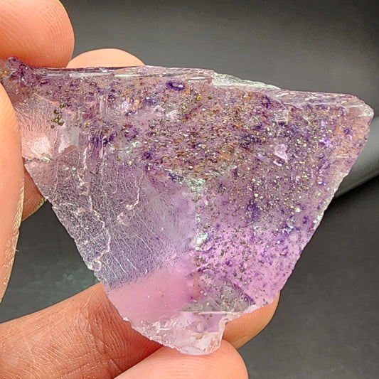 Close-up of a hand holding The Crystalary's AUCTION Fluorite, sourced from the Cave-In-Rock District, Hardin County, Illinois, USA. The crystalline fluorite features a predominantly purple hue with a partially translucent surface adorned with specks of darker purple and reflective pyrite inclusions, set against a dark background.