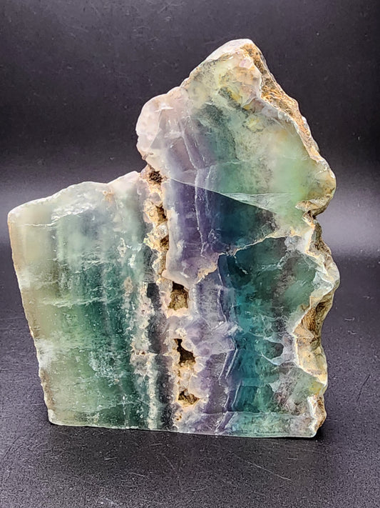 A rough and semi-polished slab of AUCTION Fluorite from Chihuahua, Mexico, displaying bands of green, purple, and blue colors. The edges are jagged and the surface shows natural striations and textures against a dark background. Brought to you by The Crystalary.