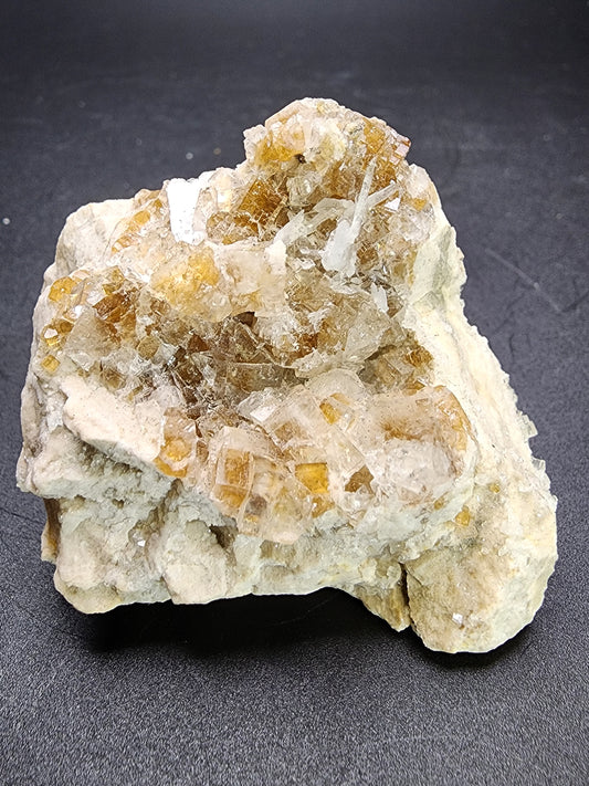 A close-up image of a mineral specimen from The Crystalary showcases translucent, yellowish-brown cubic crystals embedded in a beige, rocky matrix. The crystals vary in size and cluster on the surface, exhibiting a shiny, glass-like appearance characteristic of the Fluorite specimen from AUCTION Stoneco White Rock Quarry, Clay Center, Allen Township, Ottowa County, Ohio, USA. The background is a dark, smooth surface.