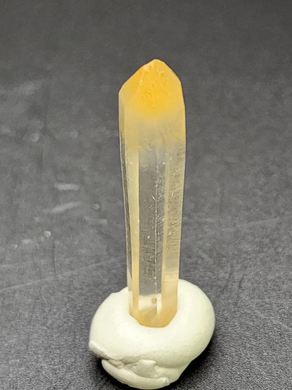 A single translucent AUCTION Mango Quartz crystal from Cabiche, Municipal de Quipama, Boyaca Dept., Columbia is standing upright, supported by a blob of white adhesive material on a dark surface. The yellowish-tinted crystal from The Crystalary has smooth, well-defined edges and a pointed tip.