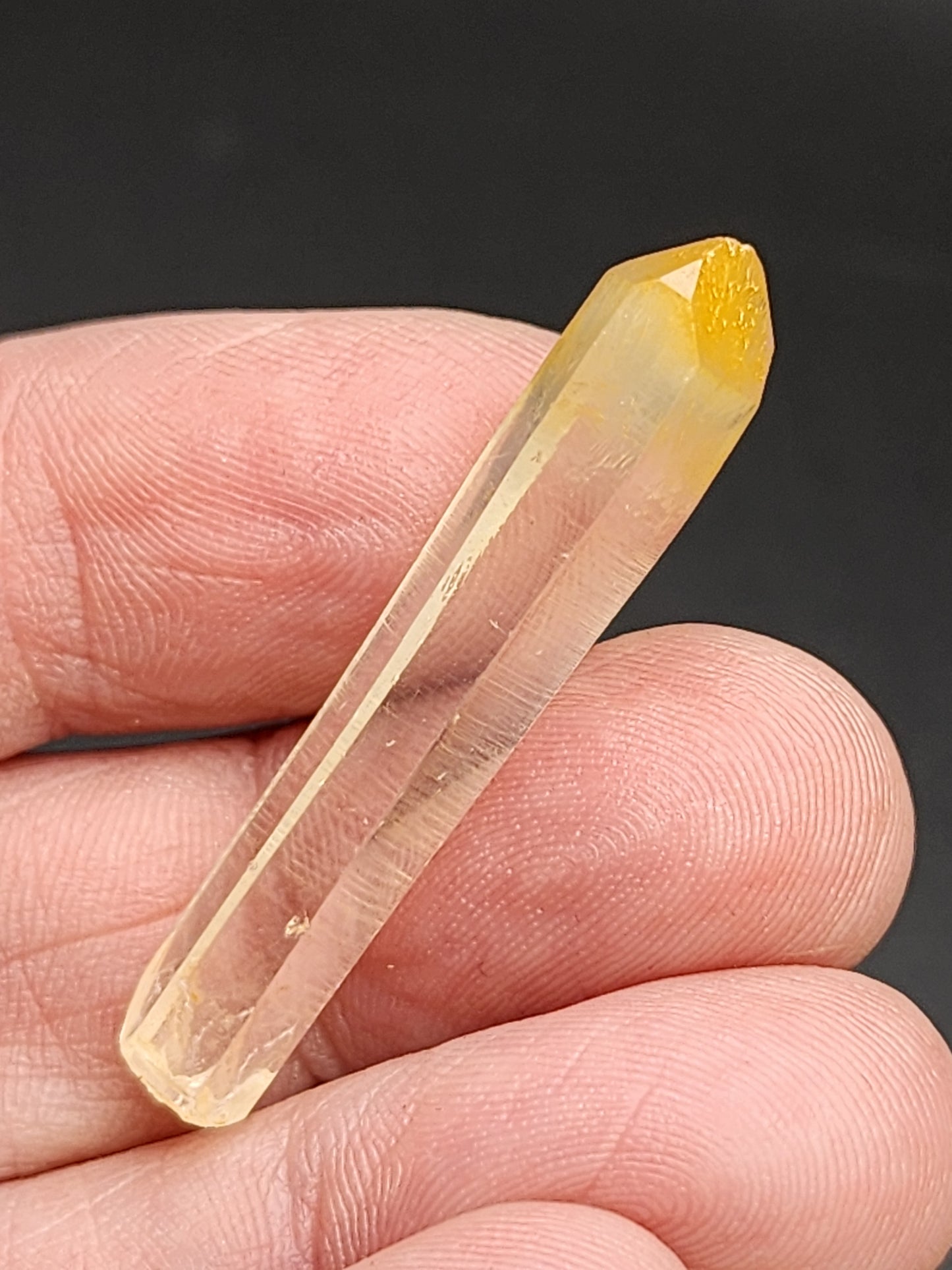 A hand holding a transparent, yellow-tinted AUCTION Quartz with Halloysite (Mango Quartz)-Cabiche from The Crystalary, featuring a pointed end and clear facets in a smooth, polished appearance. The dark background accentuates the crystal's remarkable clarity and vibrant color.
