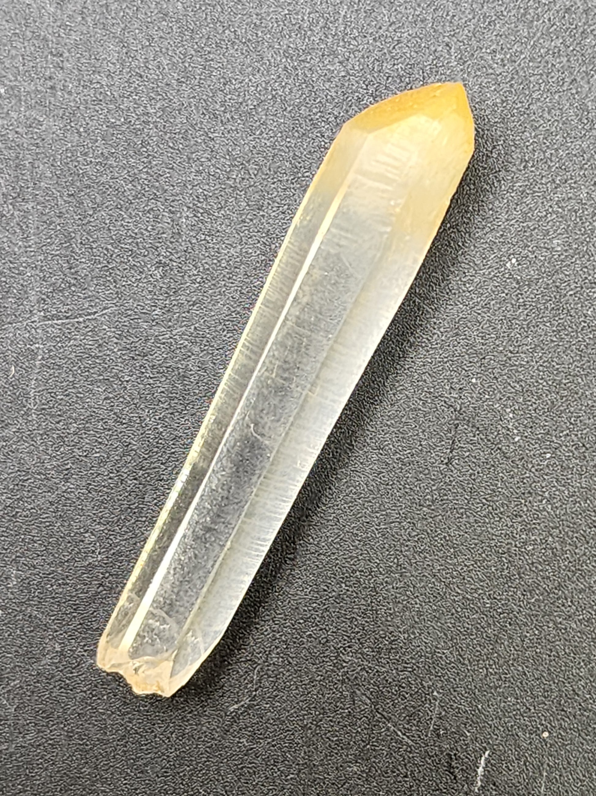 A yellow, elongated Mango Quartz crystal from The Crystalary's collection, identified as "AUCTION Quartz with Halloysite (Mango Quartz) - Cabiche, Municipal de Quipama, Boyaca Dept., Columbia," features smooth, facet-like surfaces and a pointed end. It is displayed against a textured black background. The crystal showcases a subtle gradient with a deeper yellow hue at the tip.